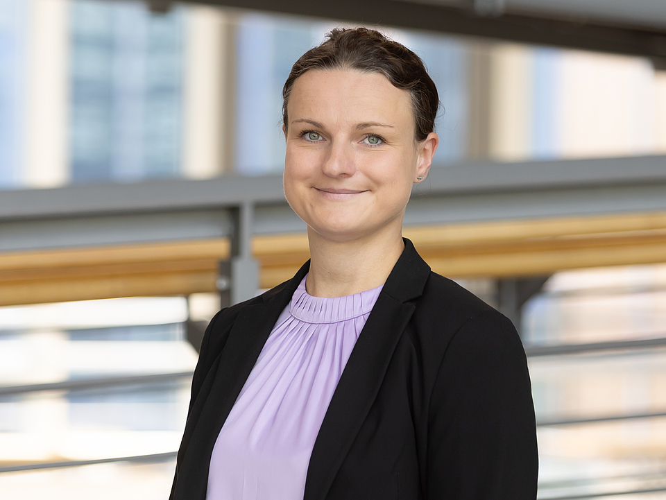  Eine junge Frau mit zusammengebundenen, dunklen Haaren und einem leichten Lächeln trägt eine lilafarbene Bluse und einen schwarzen Blazer. Sie steht im Foyer des Krankenhauses mit hellen Geländern und Fenstern im Hintergrund.