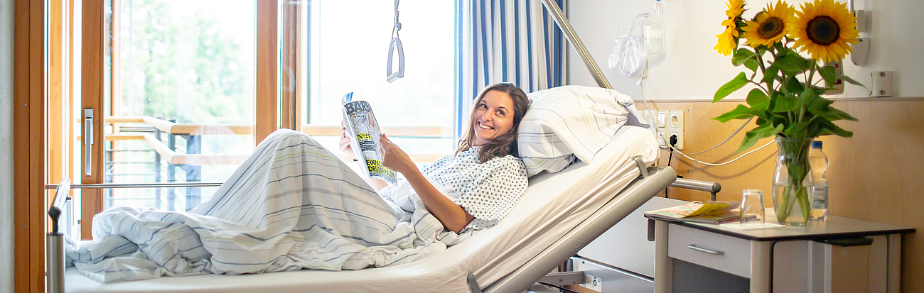 Eine Patienten liegt im Krankenhausbett mit einer Zeitschrift und lächelt in die Kamera.