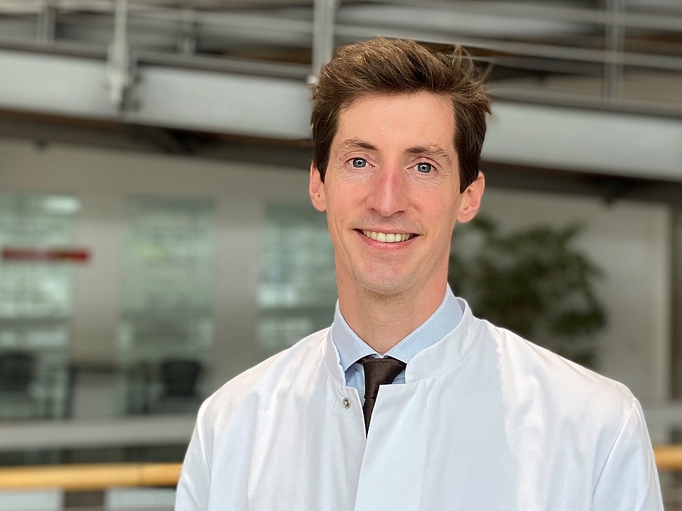 Porträt Götte Ein junger Mann mit kurzen, dunklen Haaren und einem weißen Arztkittel lächelt in die Kamera. Er steht in einem hellen, modernen Gebäude mit einer Rampe und Geländern im Hintergrund.