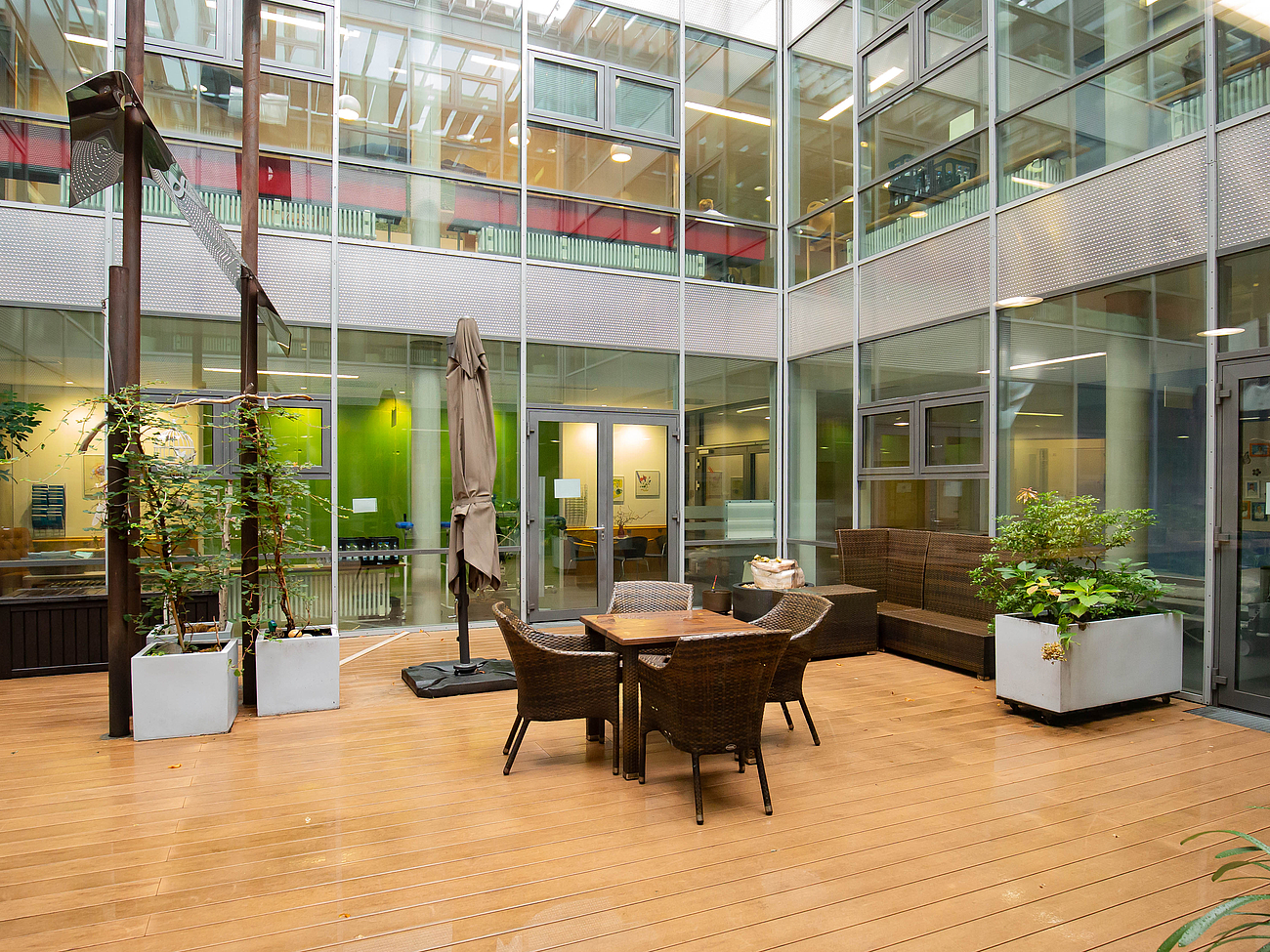 Innenhof / Palliativgarten im Krankenhaus Agatharied Ein heller, begrünter Innenhof mit Glasfassaden des Krankenhauses Agatharied, eingerichtet mit bequemen Korbmöbeln und einem Sonnenschirm. Auf dem Holzboden stehen mehrere große Pflanzkübel mit grünen Pflanzen. Die umliegenden Glasfronten reflektieren das Tageslicht.