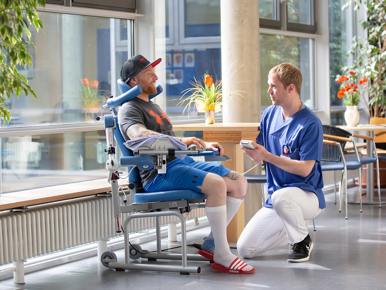 Ein Patient sitz auf einem Stuhl im Flur und vor ihm kniet ein Pfleger und hilft ihm.