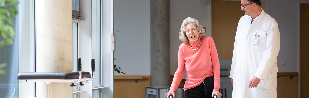 Ein Arzt in weißem Kittel begleitet eine ältere Patientin in rosa Oberteil und schwarzer Hose, die sich auf einen Rollator stützt und lächelnd zur Seite blickt. Sie gehen gemeinsam durch einen hellen Flur mit großen Fenstern.