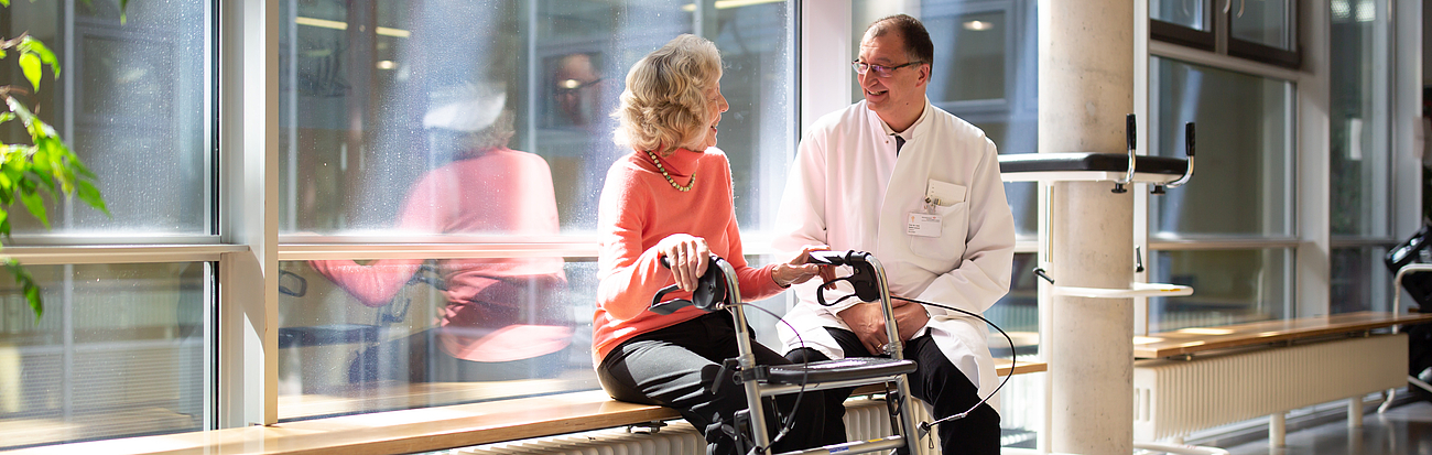  Ein Arzt in weißem Kittel sitzt auf einer Bank neben einer älteren Patientin, die ebenfalls auf der Bank sitzt und einen Rollator vor sich hat. Sie unterhalten sich in einem hellen Flur mit großen Fenstern.
