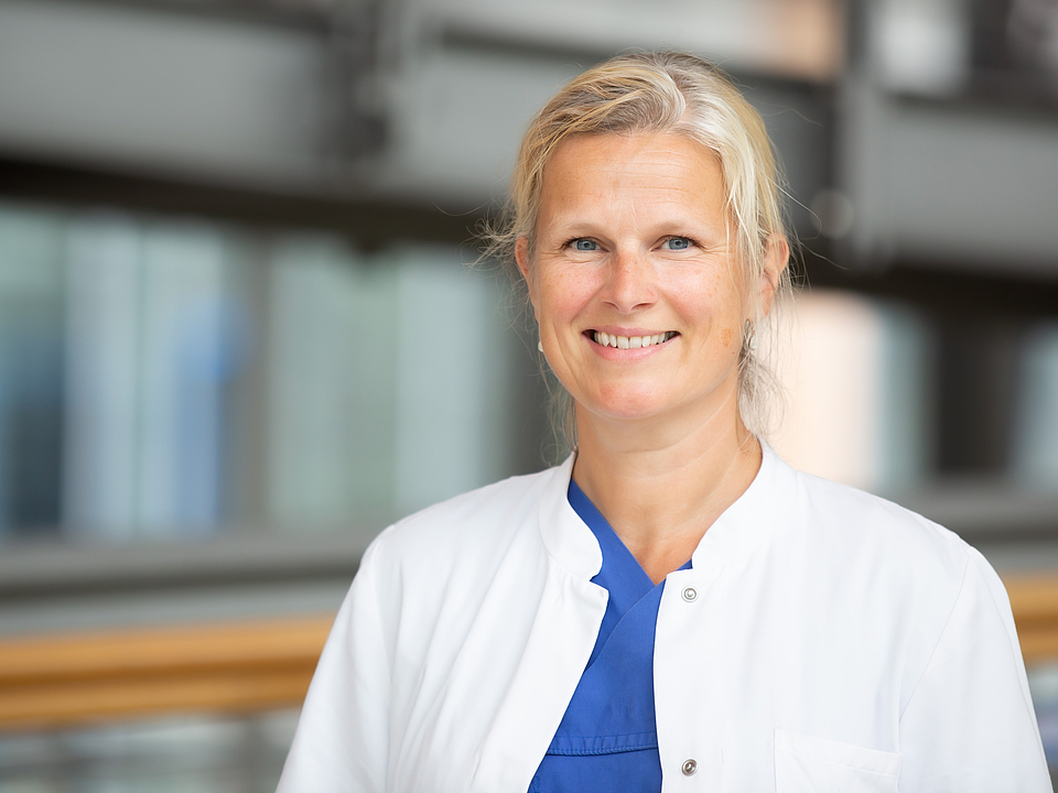 Porträt von Frau Philippson Eine lächelnde Frau mittleren Alters mit blondem Haar, die einen weißen Arztkittel über blauer medizinischer Kleidung trägt und in die Kamera blickt.