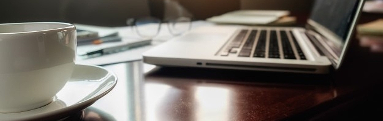 Ein Laptop und eine Tasse stehen auf einem Tisch und im Hintergrund sitzt eine Frau.